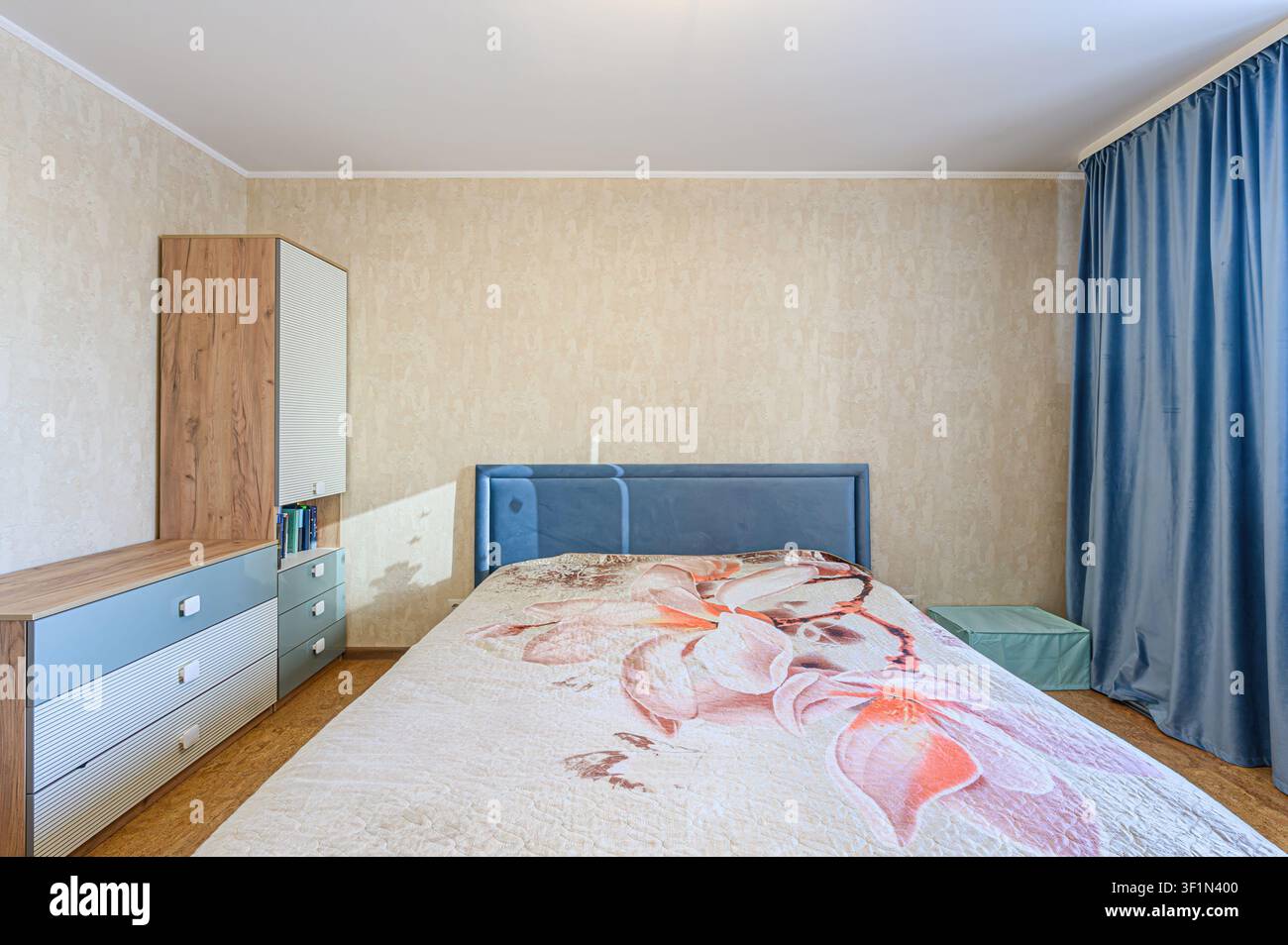 Camera da letto accogliente con un letto con trapunta floreale, una testiera blu, un armadio e tende blu. La stanza ha pareti beige e un'atmosfera chiara e calda Foto Stock