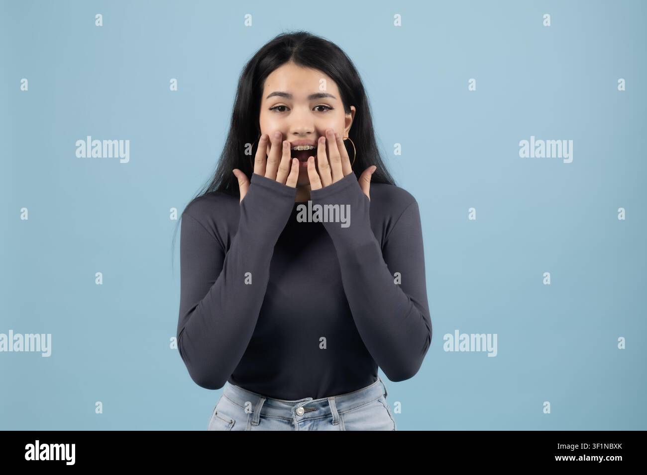 Giovane donna sorpresa che guarda la telecamera con le mani sul viso in studio Foto Stock
