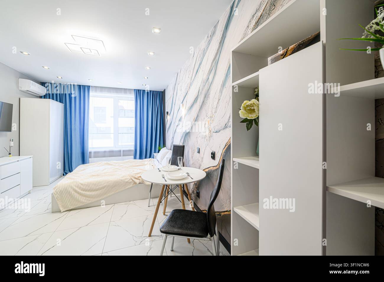 Apartamento studio luminoso con pavimenti in marmo bianco, tende blu e una parete in marmo decorativa. Sono anche visibili un piccolo tavolo da pranzo e scaffali bianchi Foto Stock