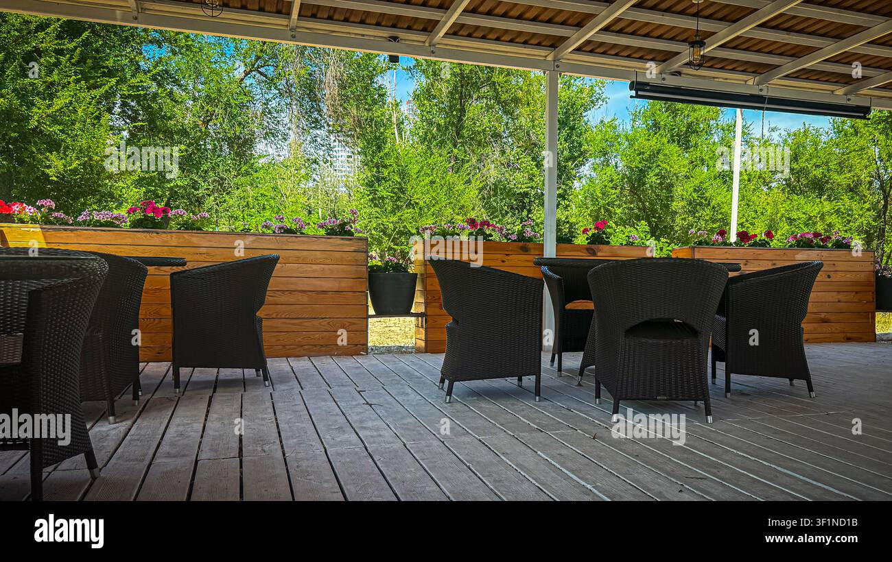 Accogliente terrazza all'aperto di un caffè con sedie di vimini scure su un ponte di legno, circondata dalla vegetazione. - Immagine stock catturata con smartphone