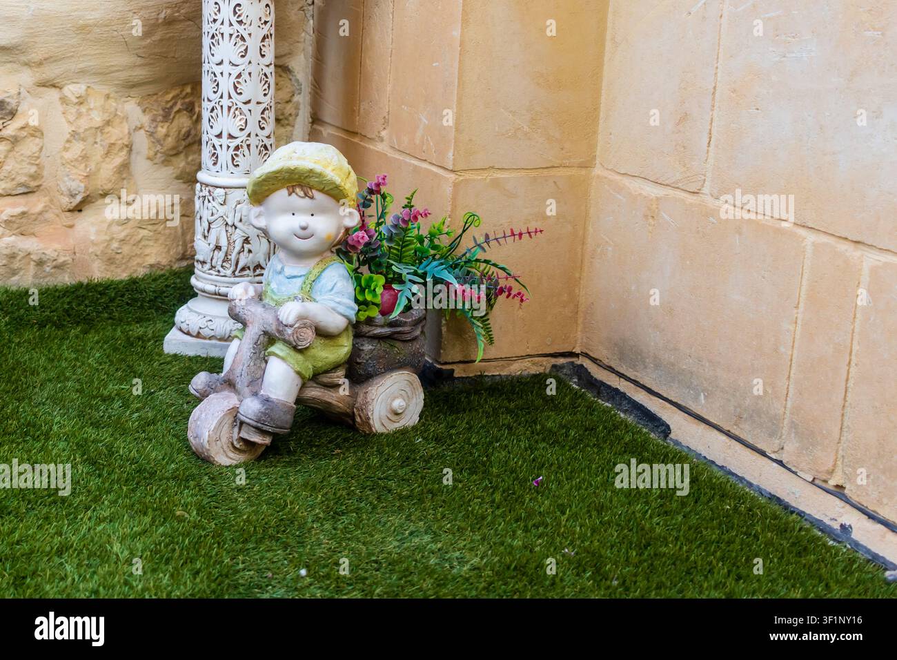 Figurina da giardino stravagante di una ragazza su un triciclo di legno con fiori contro un muro di pietra Foto Stock