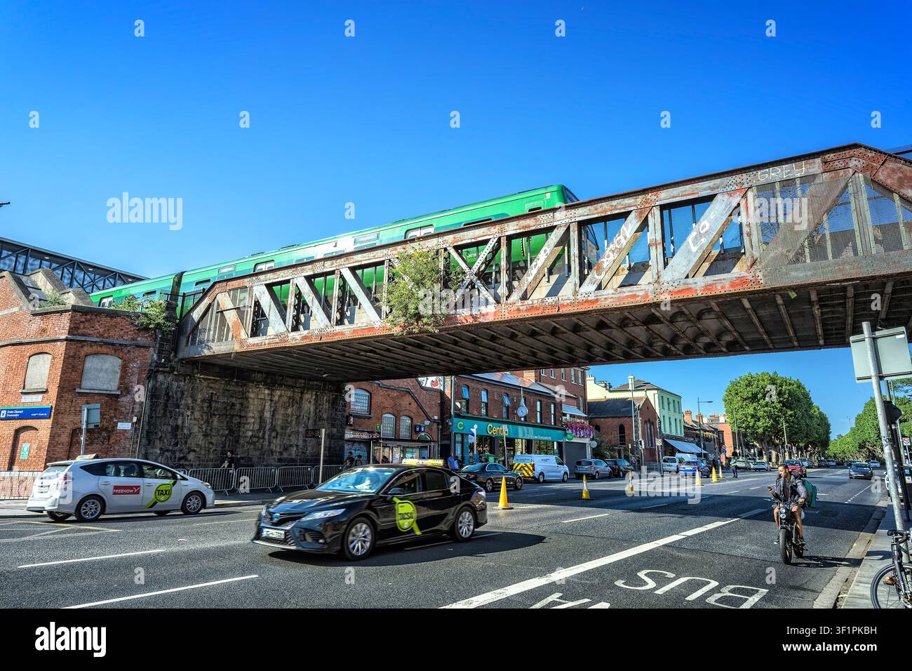 Traffico e un ciclista che passa sotto il ponte ferroviario storico in ferro arrugginito della stazione di Drumcondra a Dublino con un treno pendolare verde che lo attraversa Foto Stock