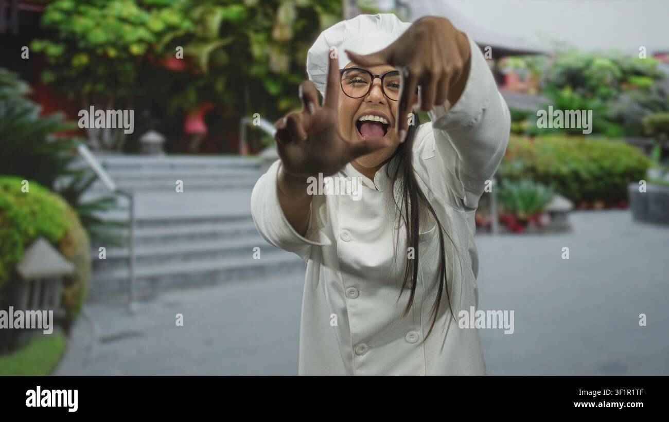 Giovane donna chef con uniforme e cappello bianchi, che incornicia la fotocamera con le mani e sorride in un'area delle scale del cortile; creatività giocosa. Foto Stock
