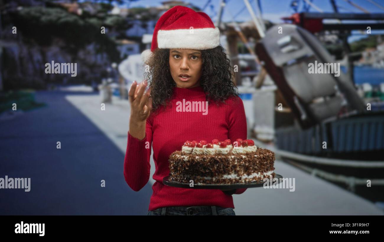 Donna con capelli ricci che indossa un cappello di Babbo Natale e un maglione rosso tiene una torta al cioccolato su un vassoio mentre tocca il suo cappello in una marina; frustrazione natalizia. Foto Stock