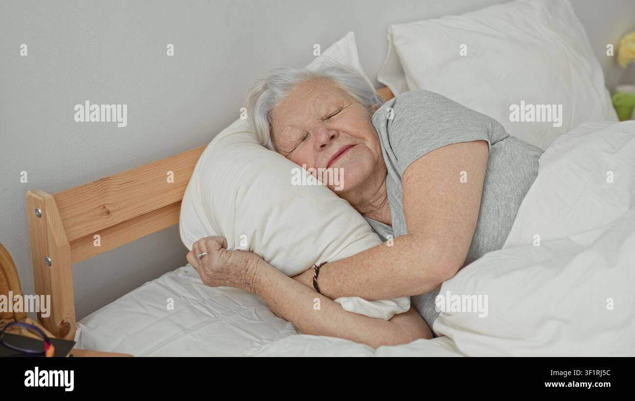 Una donna anziana abbraccia un cuscino mentre dorme su un letto in una camera da letto, distesa sul fianco con gli occhi chiusi e le mani che stringono il cuscino; comfort serenità. Foto Stock