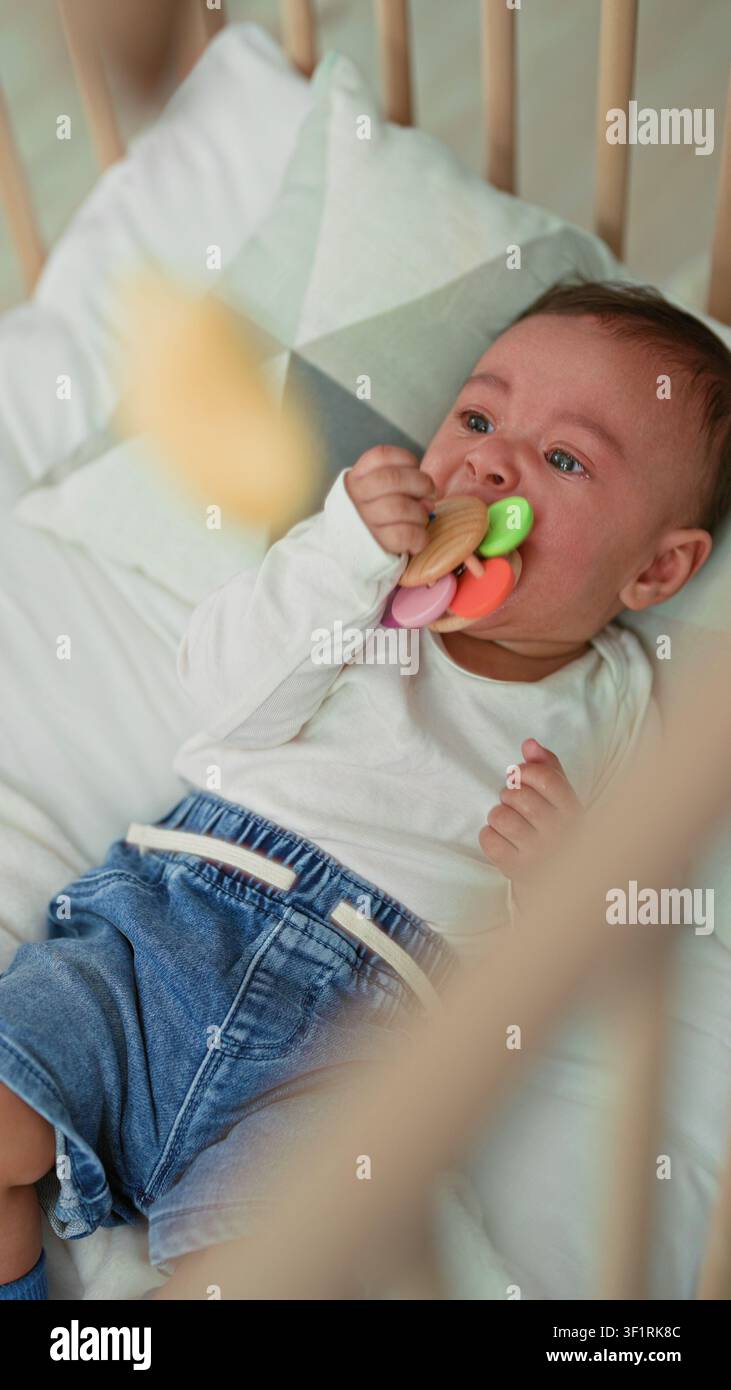 Bambino ispanico nella culla mordendo un anello colorato con bocca e occhi lacrimi in un asilo nido di un edificio; fastidio dentifricio. Foto Stock