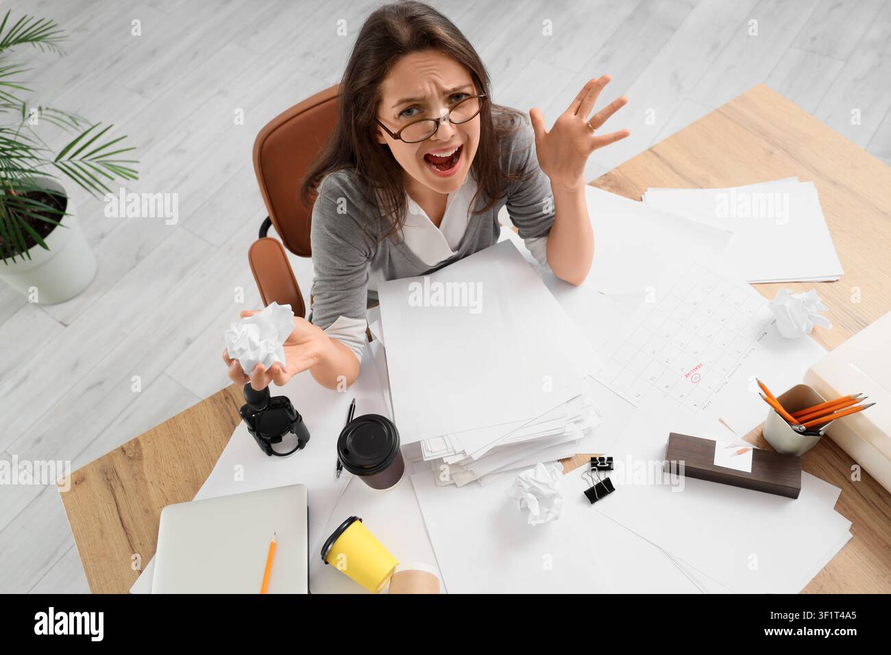Giovane donna frustrata con carta accartocciata su una scrivania disordinata, concetto di stress per scadenze e pressione lavorativa, impatto emotivo delle sfide professionali Foto Stock