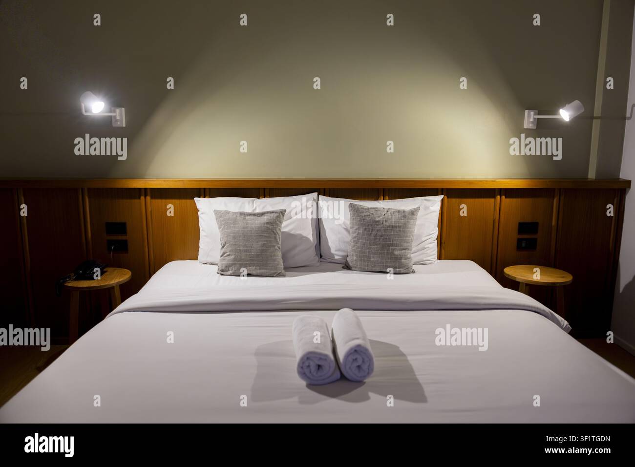 Un letto di camera d'albergo moderno con lenzuola bianche, due asciugamani arrotolati e illuminazione soffusa che crea un'atmosfera accogliente e tranquilla Foto Stock