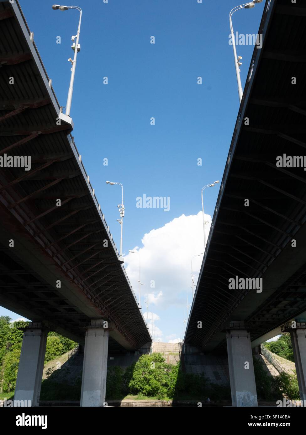 Vista verticale simmetrica che guarda verso l'alto tra due strutture parallele di ponte, lasciando un intervallo luminoso di cielo blu e nuvole. Perfetto per una g urbana astratta Foto Stock