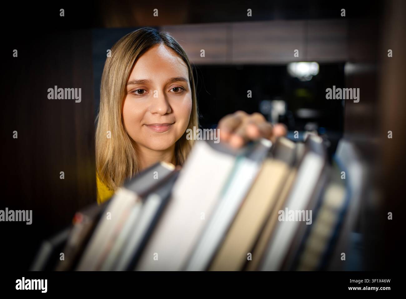 Donna che sceglie un libro da una libreria, guardando i dorsi con un sorriso gentile. Libreria o biblioteca, hobby letterario e selezione di lettura. Foto Stock
