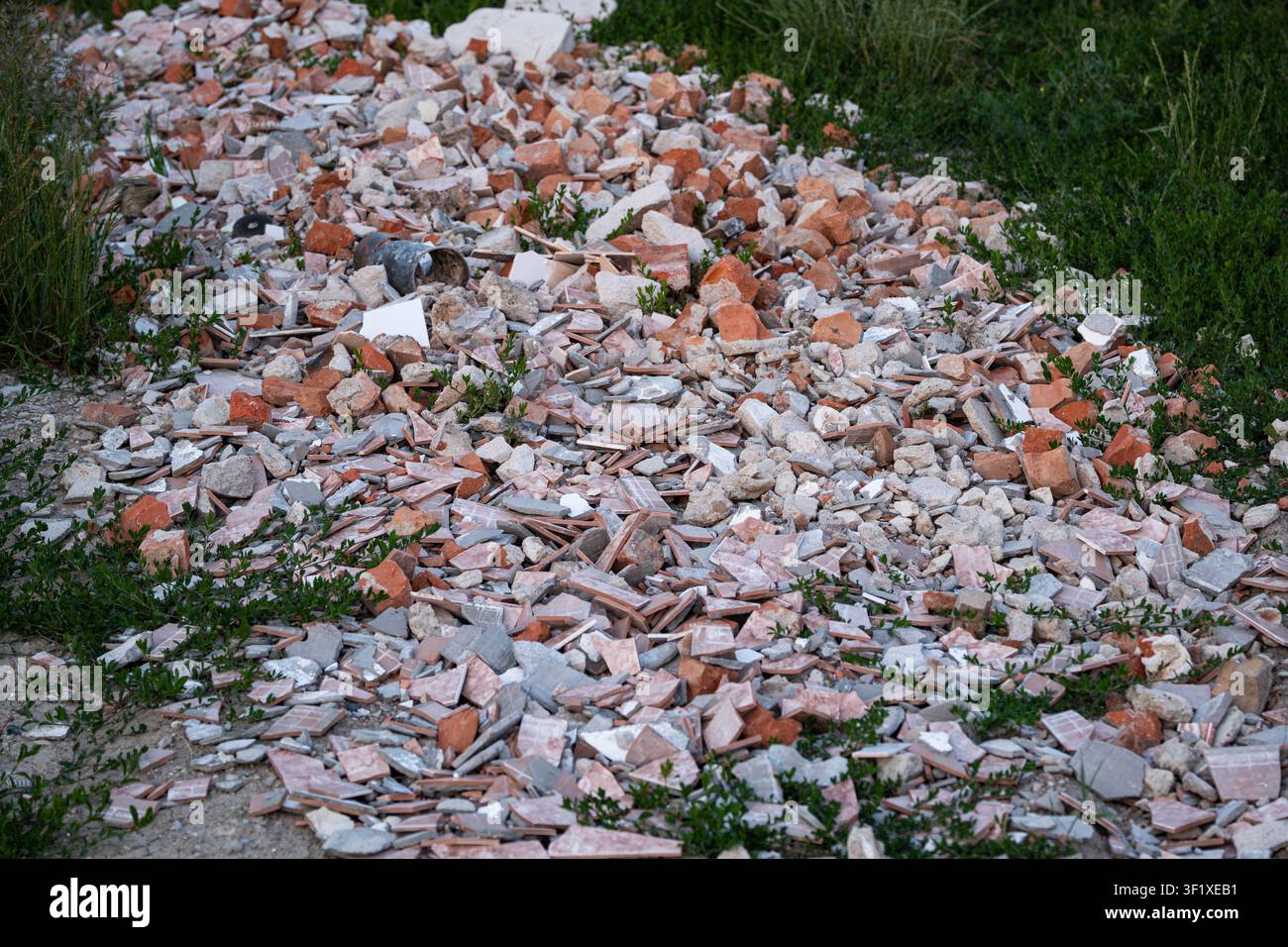 Un mucchio disordinato di rifiuti da demolizione, tra cui mattoni rossi rotti, piastrelle di ceramica frantumate e detriti di cemento, scaricati su un campo erboso all'aperto Foto Stock