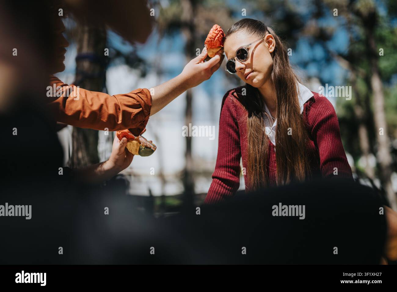 Studenti delle superiori seduti all'aperto mentre degli amici tengono giocosamente un dolce vicino alla testa di una ragazza Foto Stock