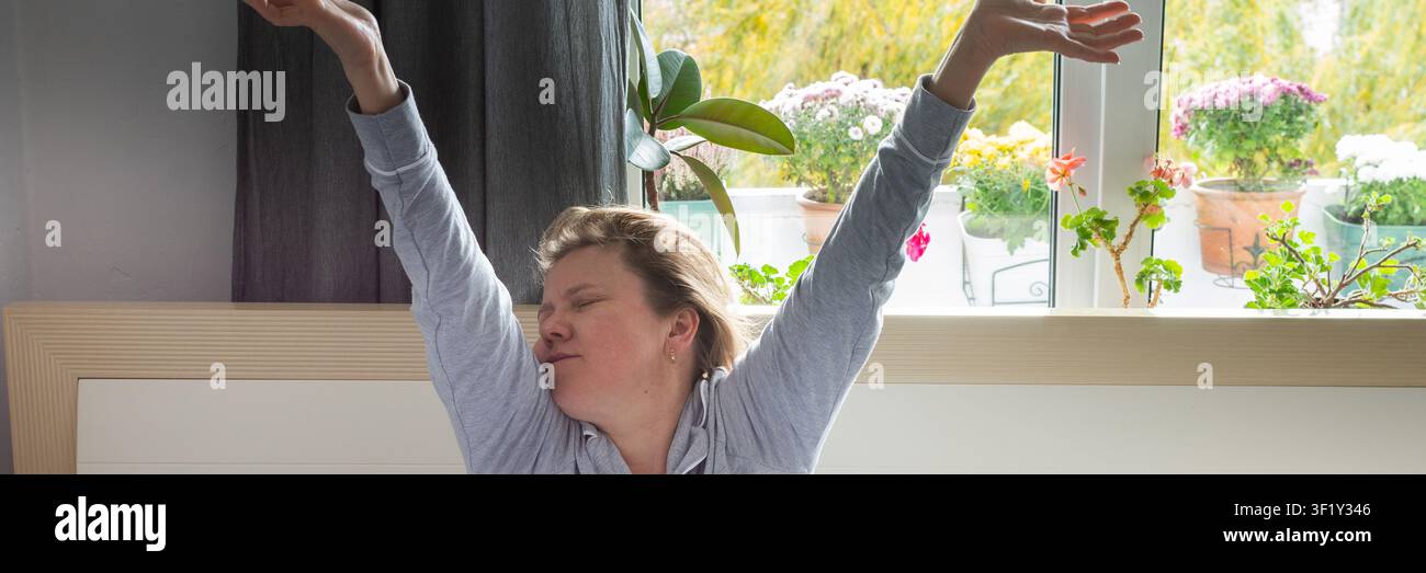 Una donna di mezza età si siede a letto, allungando le braccia sopra la testa. Sembra stanca e non ha dormito abbastanza. La sala è luminosa e naturale Foto Stock