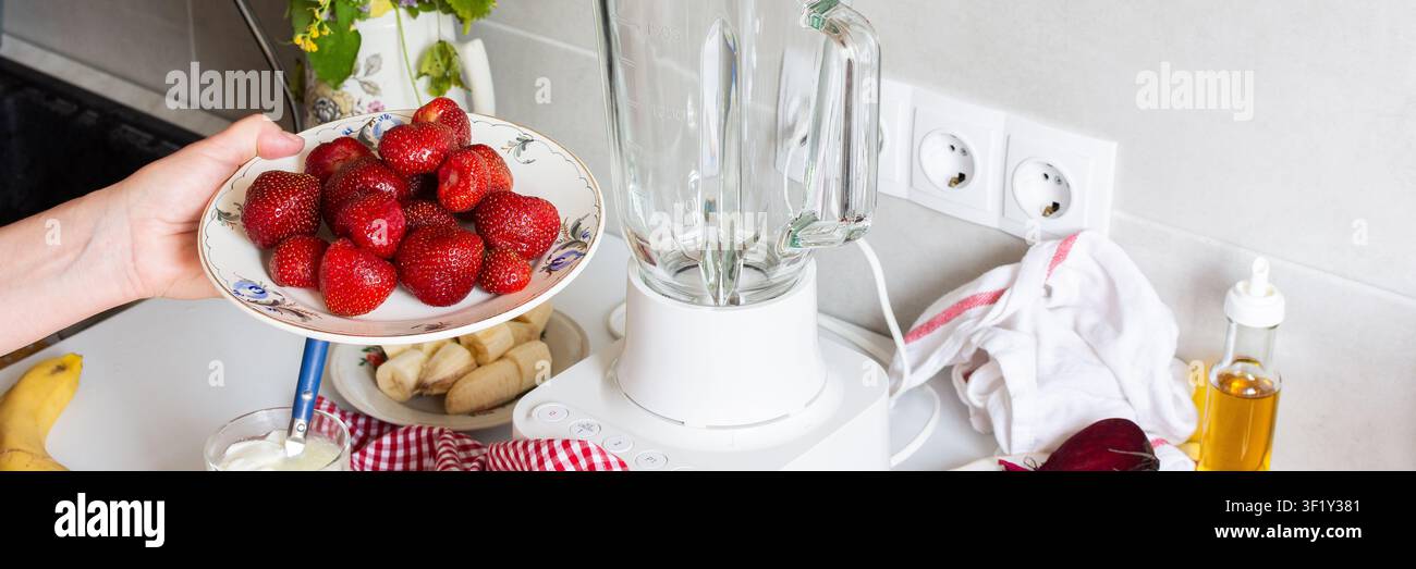 Una persona tiene un piatto di fragole fresche mentre si prepara a preparare il gelato in cucina. Ingredienti come banane e barbabietole sono anche sul conto Foto Stock