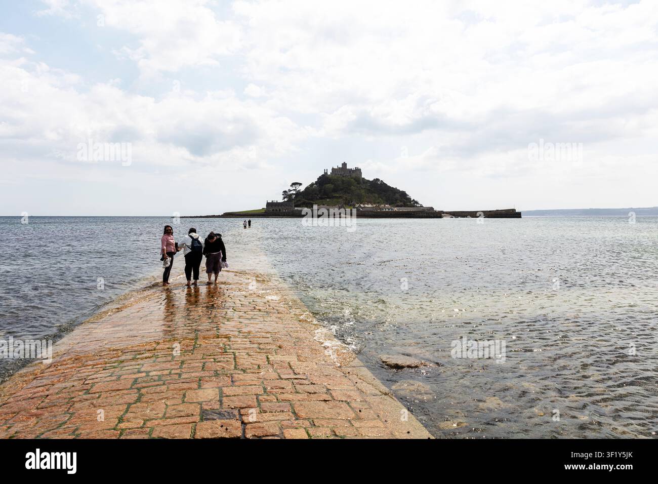 Persone che camminano lungo la diga della marea verso St Michael’s Mount in Cornovaglia. Il sentiero di pietra si estende attraverso acque basse e un bagnasciuga coperto di alghe Foto Stock
