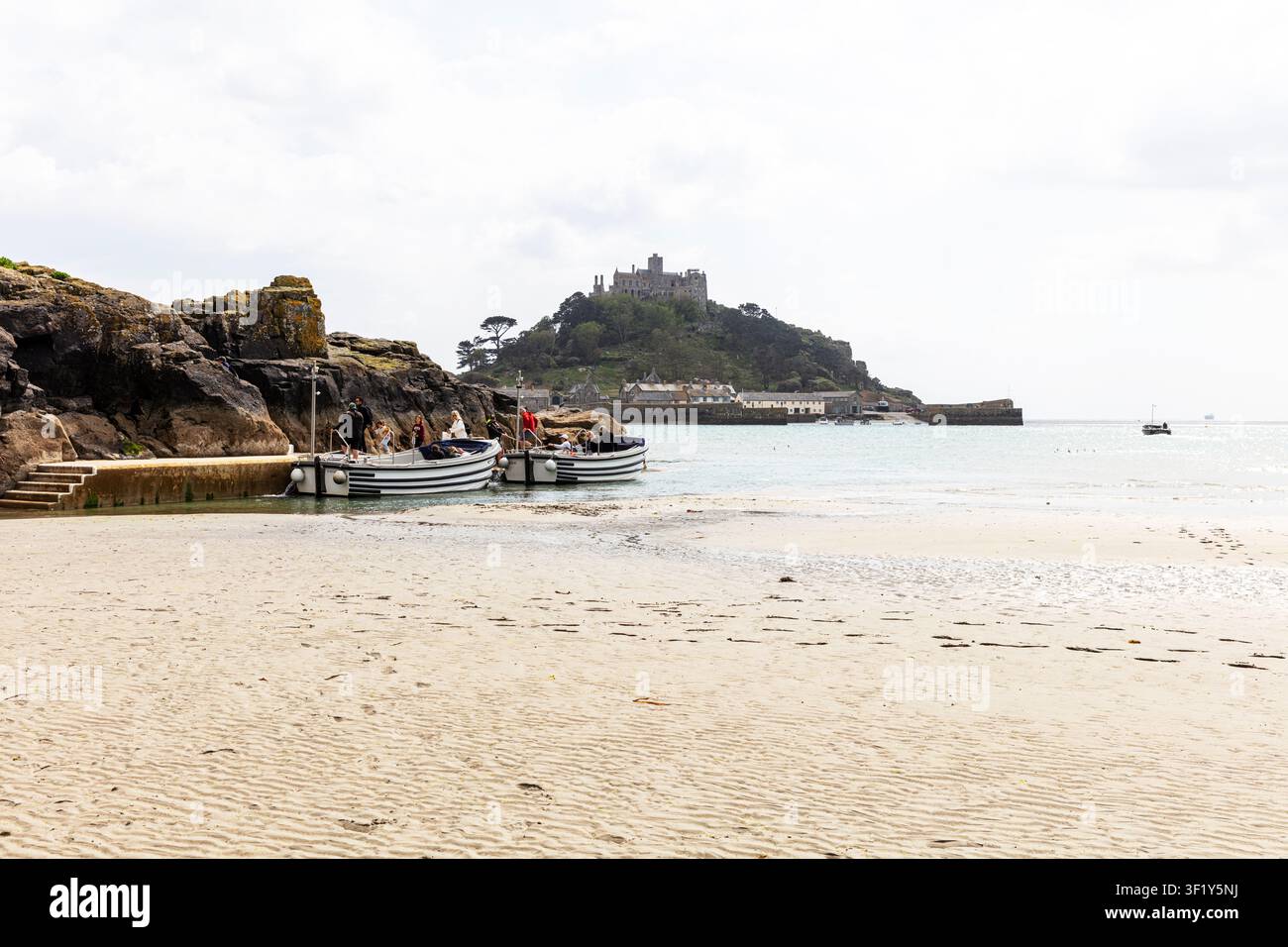 Gite in barca verso St Michael’s Mount in Cornovaglia, con spiaggia sabbiosa e piccole barche che riposano vicino alla riva. Acque calme conducono verso l'isola Foto Stock