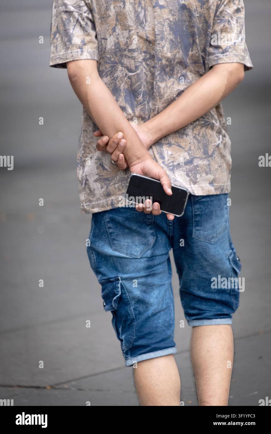 Una persona non identificata che tiene il cellulare dietro la schiena mentre cammina per una strada nel quartiere di Queens a New York. Foto Stock