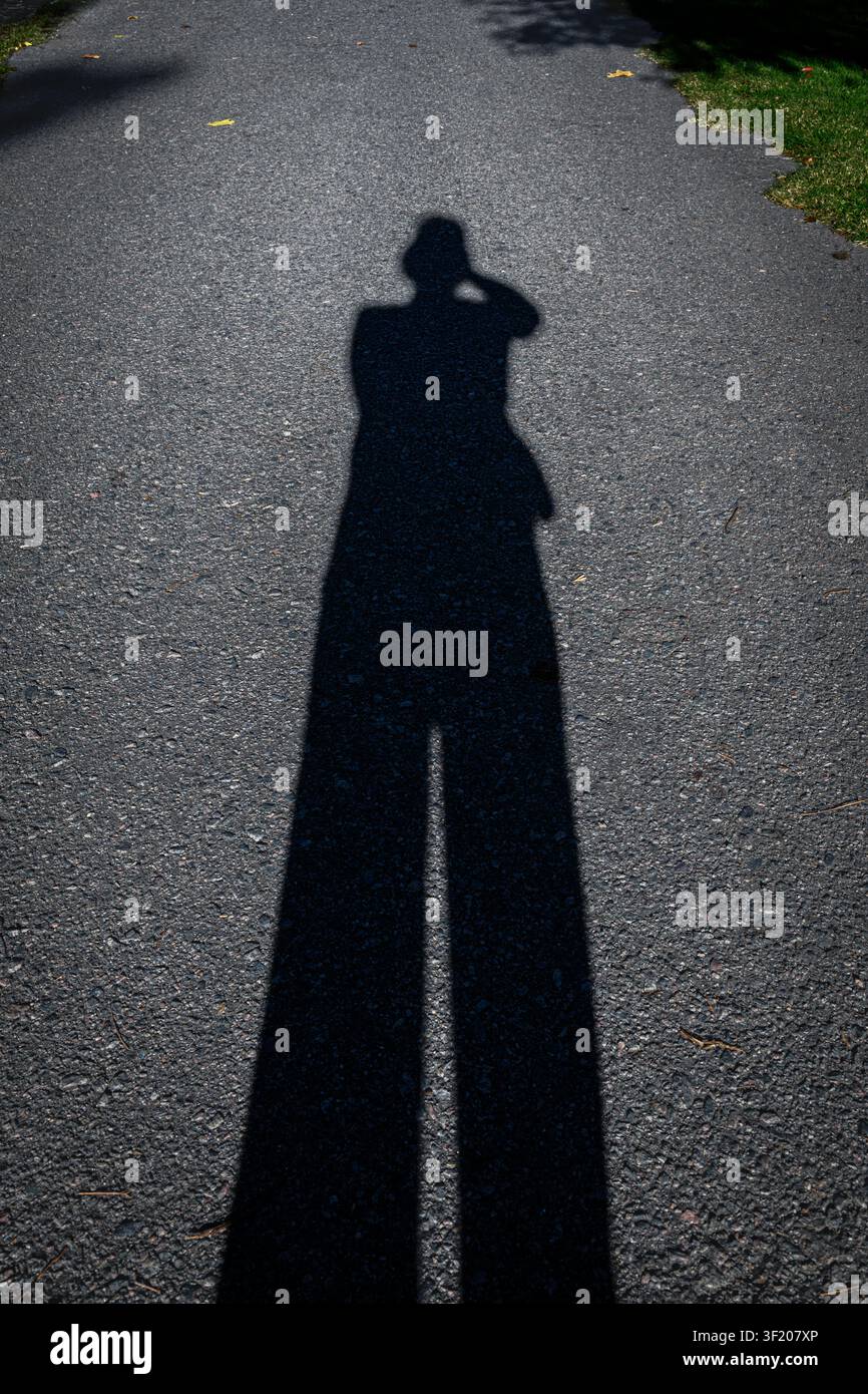 Ombra di una persona si staglia su un sentiero pavimentato. Una lunga silhouette si protende verso il cielo, quieta e profonda. Mano sollevata vicino alla testa, come a proteggere gli occhi. Bordo dell'erba Foto Stock