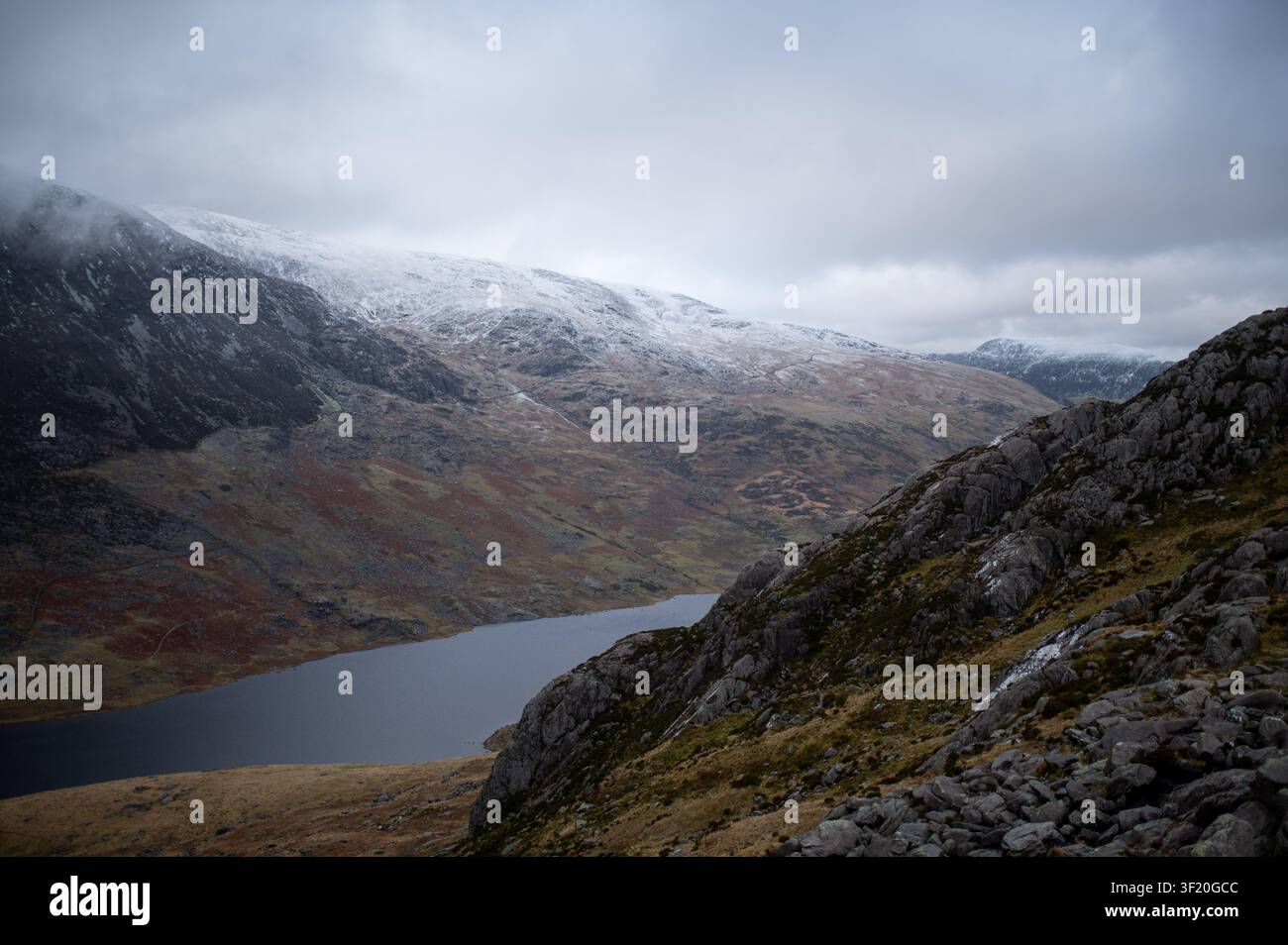 Picchi innevati incorniciano un lago tranquillo e profondo. Cieli grigi premono bassi su pendici frastagliate e muschiose. Il gelo sussurra lungo i bordi frastagliati, accennando al freddo. Immobile Foto Stock