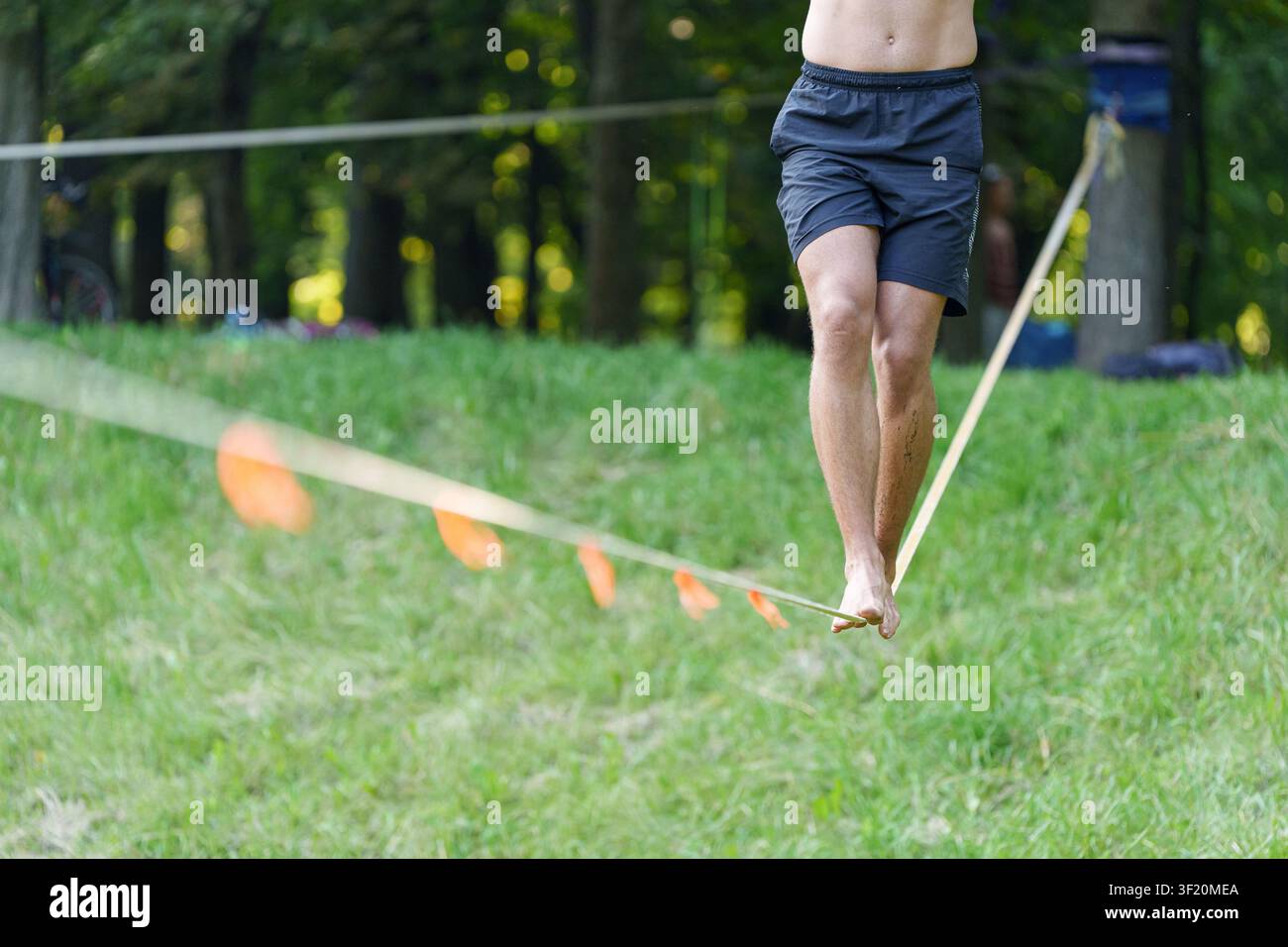 Uomo scalzo che cammina passo dopo passo su una slackline attraverso un parco verde, un piede davanti all'altro – concetto di perseveranza ed equilibrio nella vita. Foto Stock