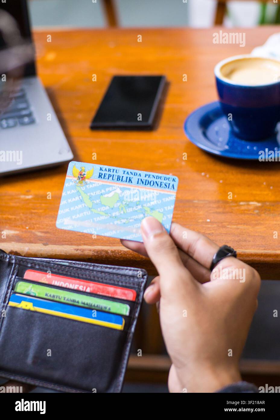 Primo piano di una persona che tiene una carta d'identità indonesiana (KTP) accanto a un portafoglio e un laptop in un caffè, verifica dell'identità online. Foto Stock