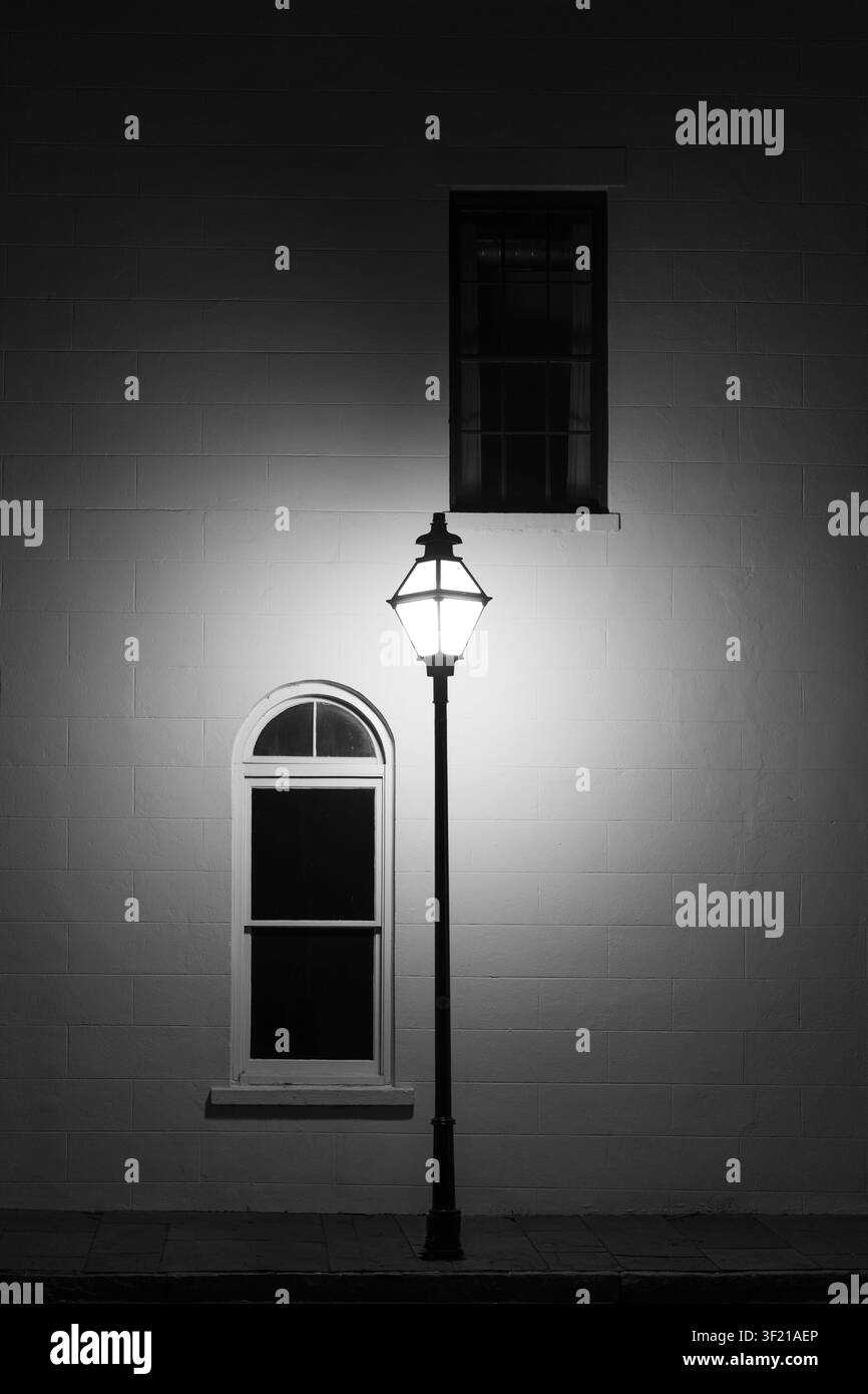 Un lampione vintage luminoso si erge accanto a due finestre su una facciata di edificio tranquilla di notte, proiettando una luce soffusa. Foto Stock