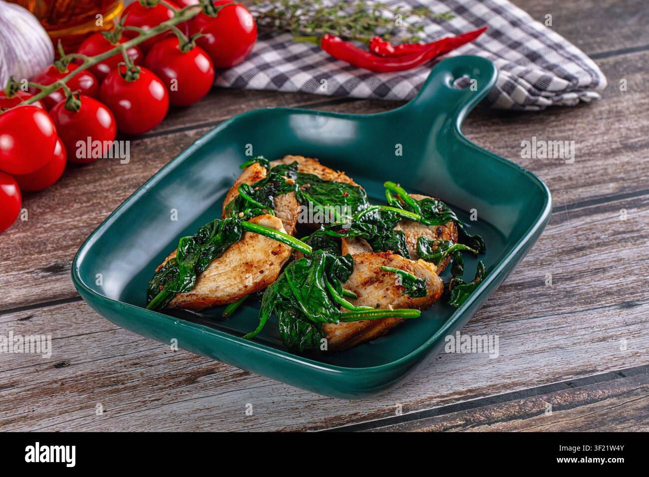 Pollo grigliato con spinaci appassiti, servito su un piatto di ceramica verdeazzurro. Lo sfondo include pomodorini freschi, aglio, peperoncino rosso e un panno a quadri Foto Stock