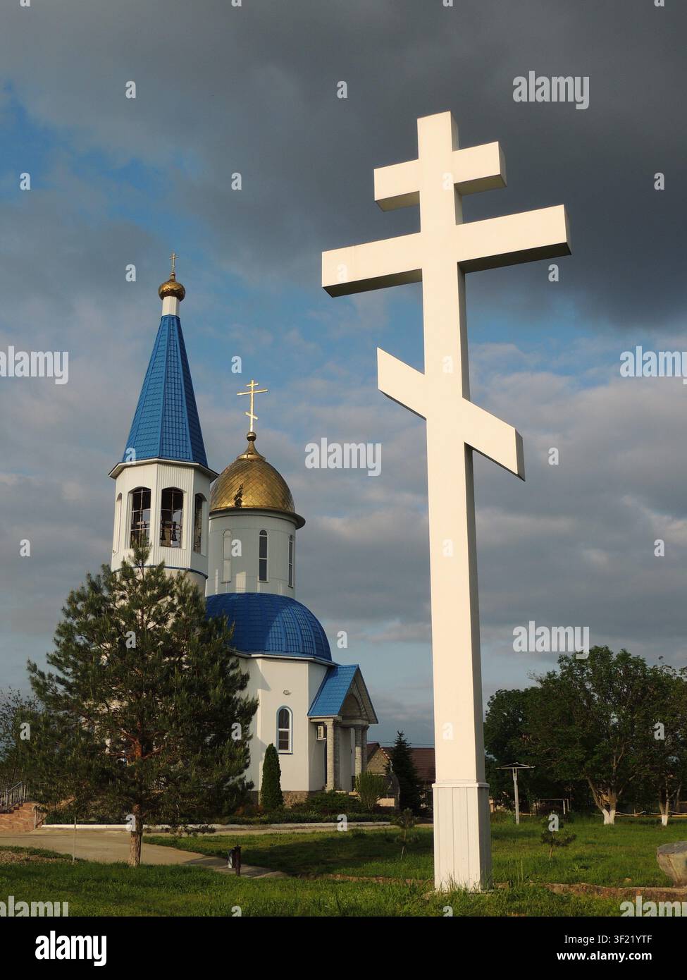 Una croce bianca monumentale si erge in primo piano. Dietro di essa è visibile una chiesa ortodossa con tetti blu, un campanile e una cupola dorata Foto Stock