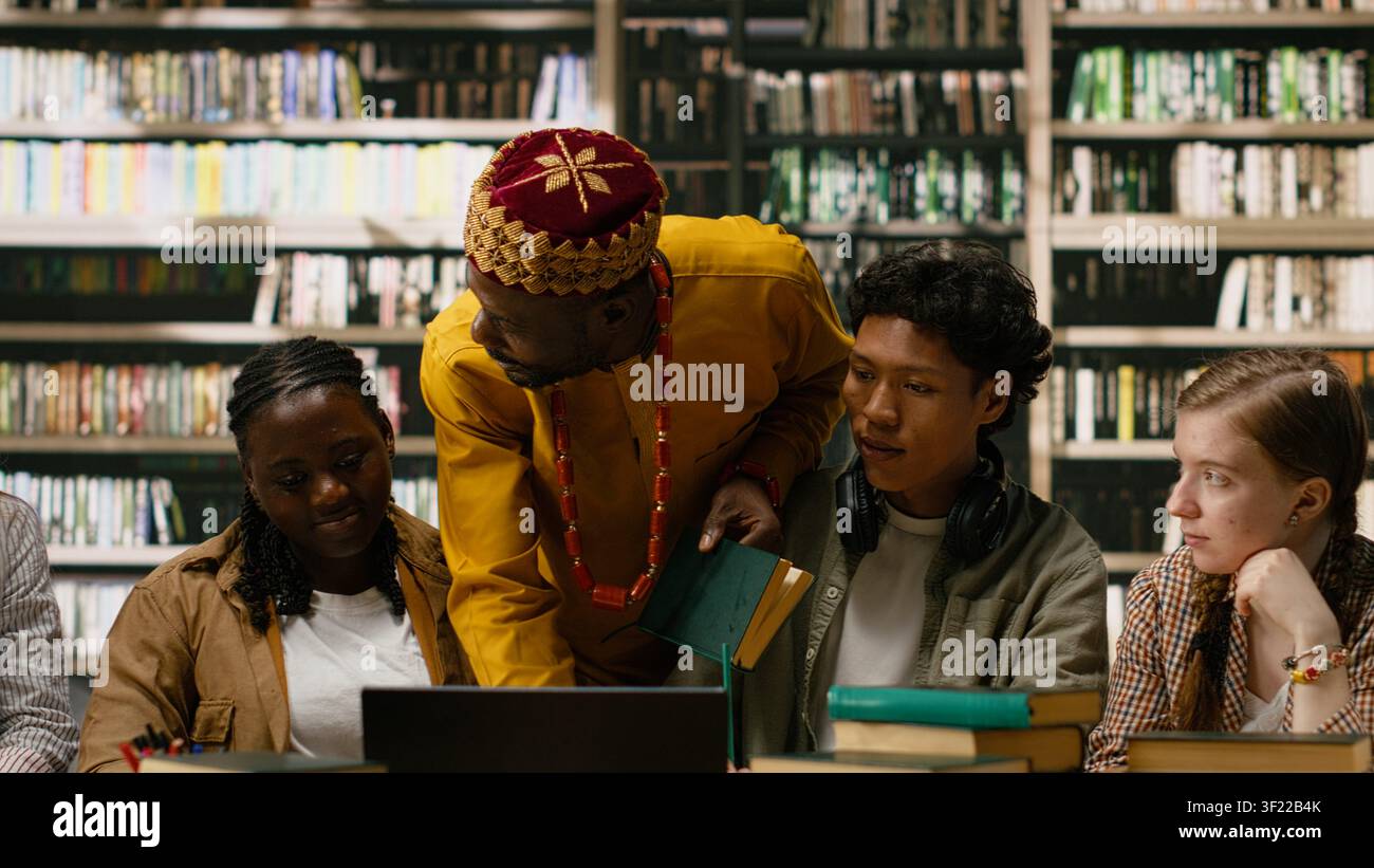 Diversi compagni di classe in studio di gruppo si riuniscono intorno a un laptop con materiale di studio su un tavolo della biblioteca, una scrivania con libri impilati. Condividono idee durante una sessione di apprendimento collaborativo. Foto Stock