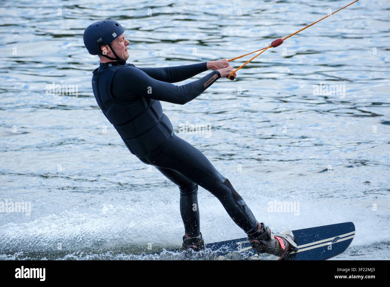 Kyiv, Ucraina - 11 luglio 2026: Un surfista su wakeboard con casco e giubbotto protettivo si piega contro la trazione del cavo mentre scivola sul Foto Stock