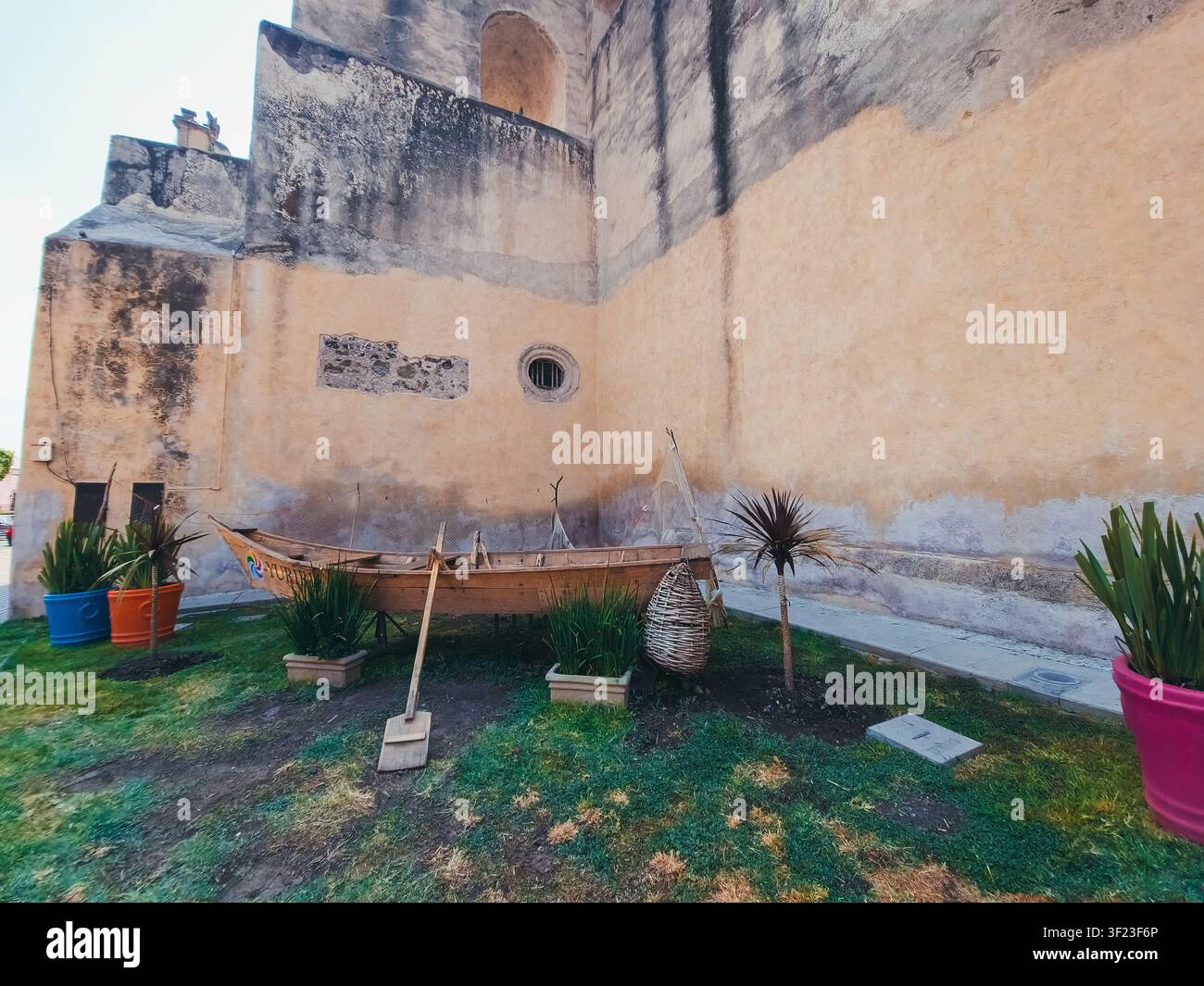 Una piccola barca è posizionata in un'area erbosa accanto a un edificio. La barca è vecchia ed è in rovina. L'area è circondata da piante in vaso, includ Foto Stock