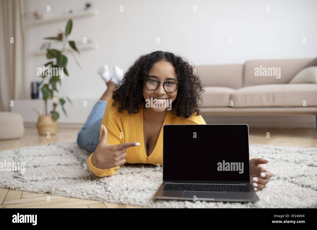 Donna afroamericana sorridente che indica lo schermo del laptop mentre è sdraiata su un tappeto a casa Foto Stock
