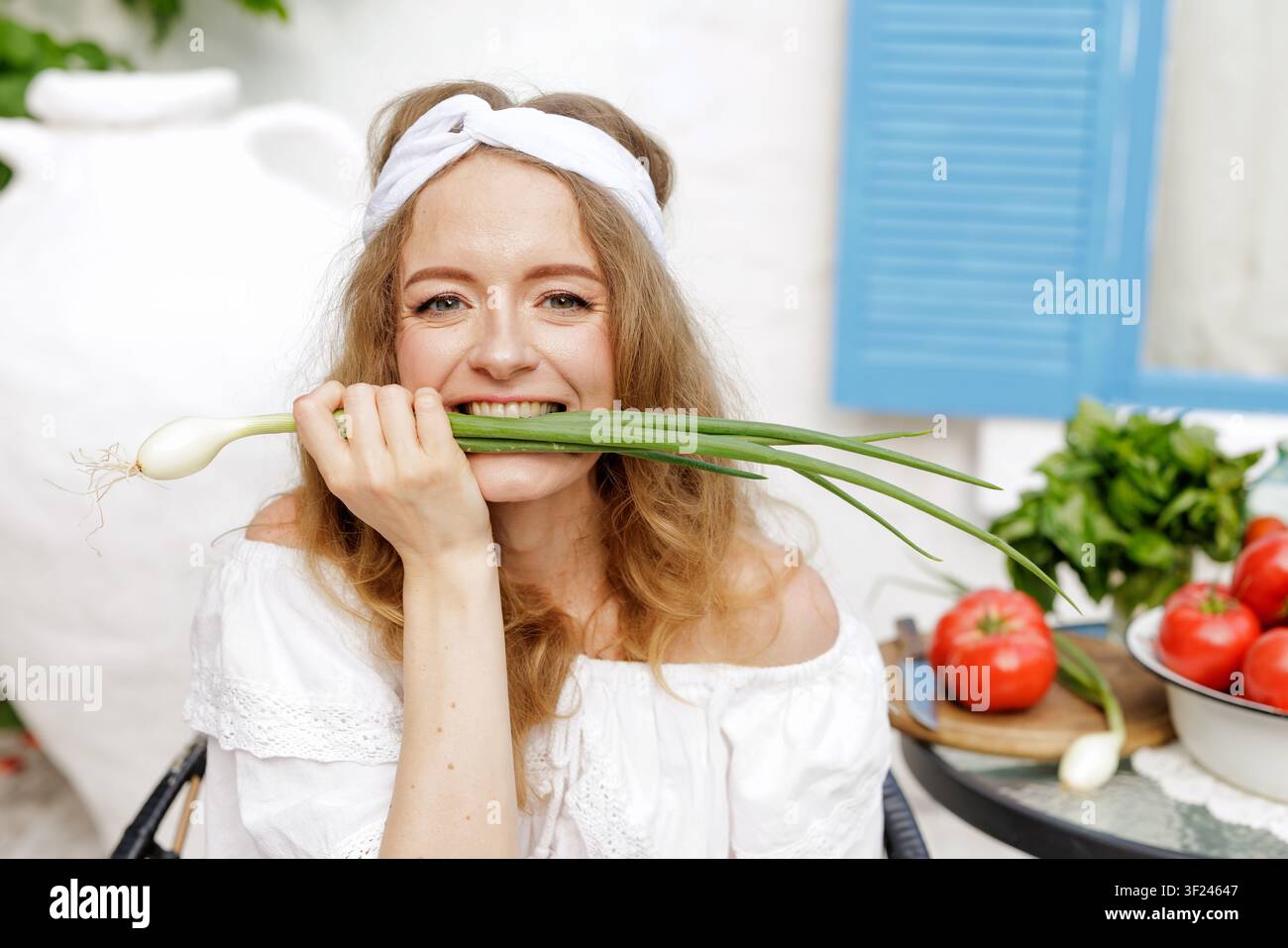 Una donna allegra morde giocosamente cipolle verdi fresche circondata da pomodori e erbe vibranti, perfetta per un tema di cucina sana. Foto Stock