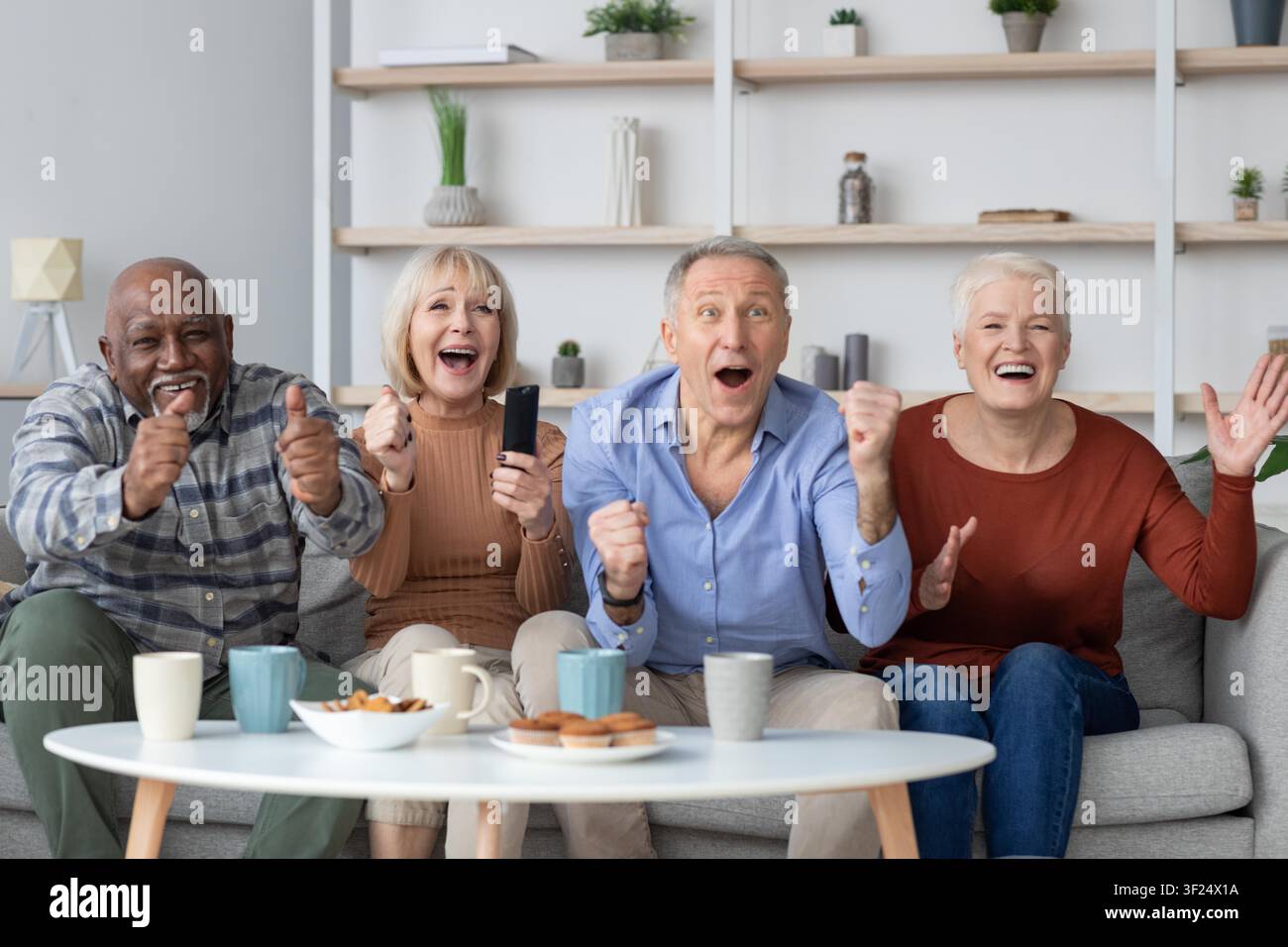 Anziani multirazziali felici che esultano insieme guardando la TV a casa, godendosi il tempo in famiglia Foto Stock
