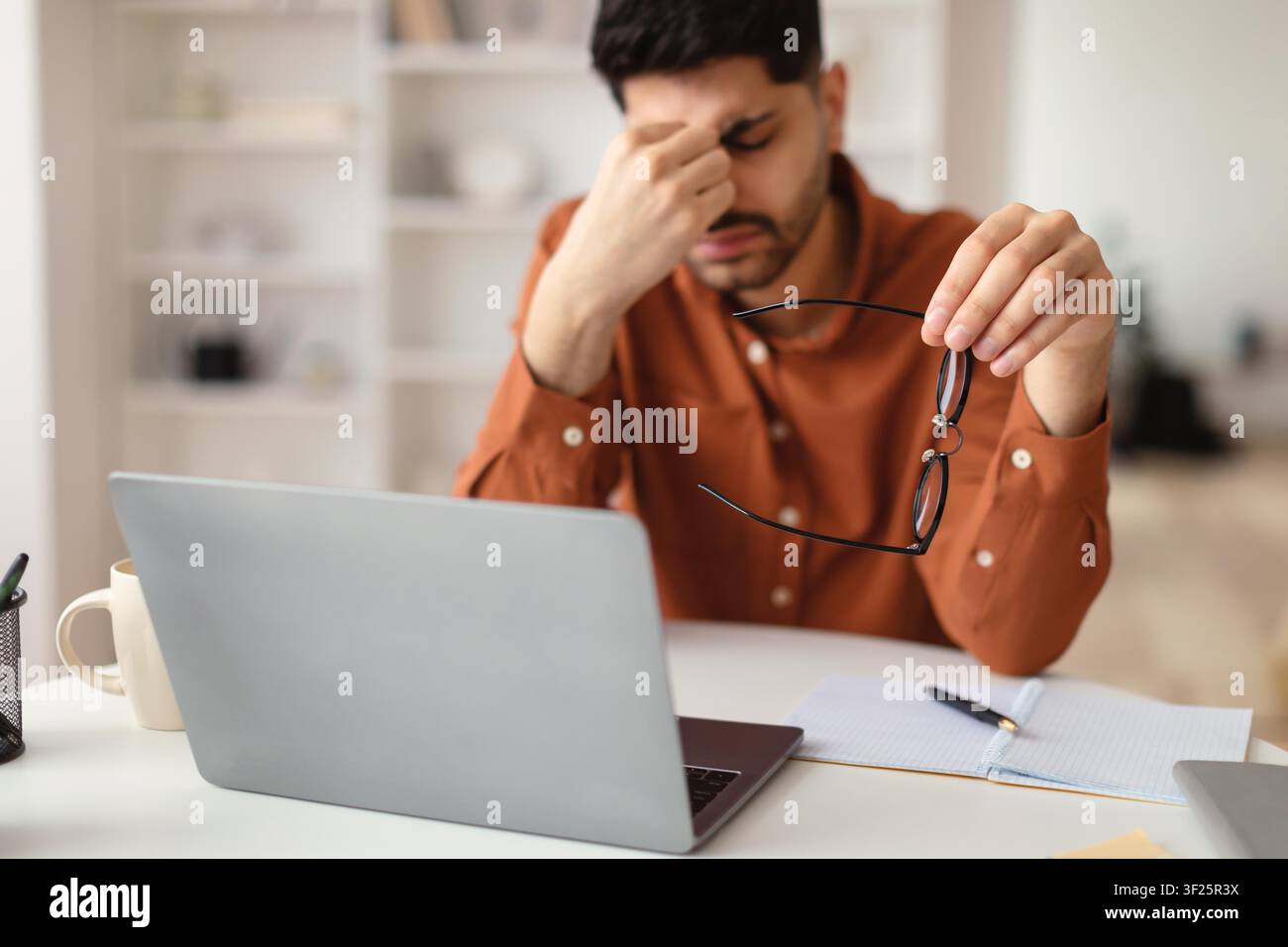 Uomo mediorientale preoccupato che tiene occhiali, stressato al computer portatile in un ufficio moderno, difficoltà lavorative Foto Stock