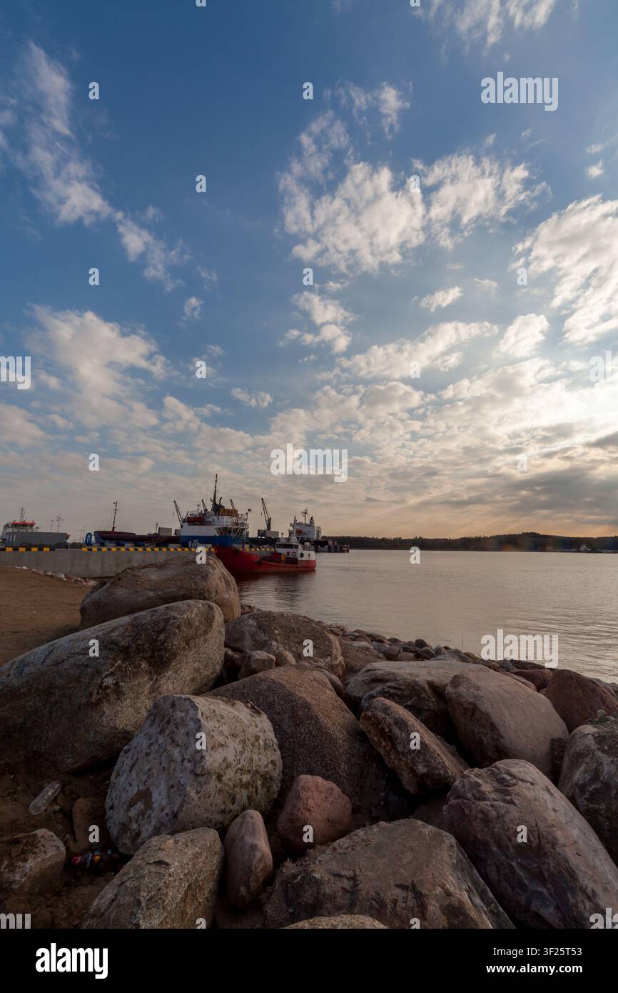 Costa rocciosa che si affaccia su un porto tranquillo con barche e un drammatico cielo nuvoloso al tramonto sopra l'acqua immobile. Foto Stock