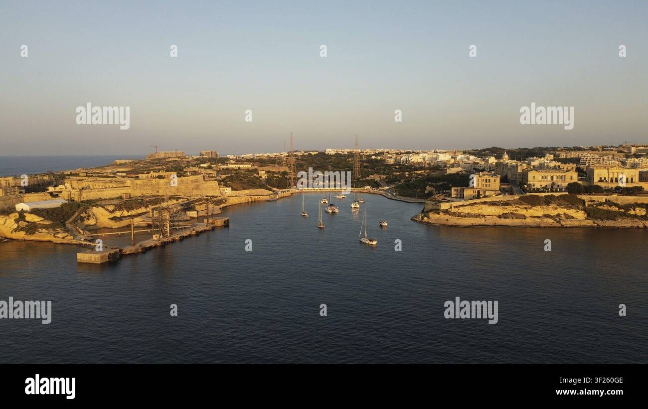 Tranquilla scena del porto con barche ed edifici lungo la costa al tramonto. Baia di Rinella, Malta Foto Stock