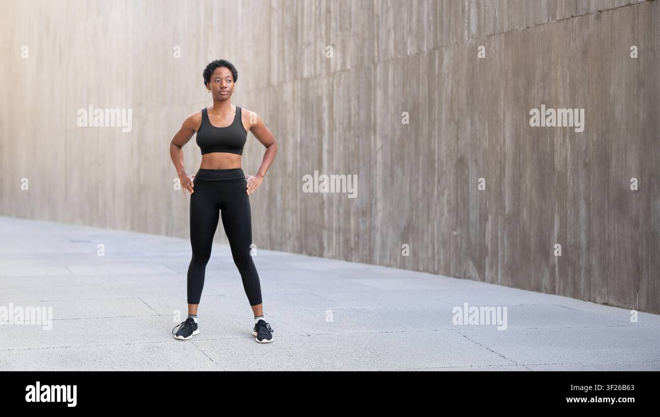 Donna nera con capelli corti che indossa abbigliamento sportivo nero e scarpe da ginnastica nere, in piedi su un'area urbana lastricata davanti a un muro di cemento prima di un allenamento all'aperto. Foto Stock
