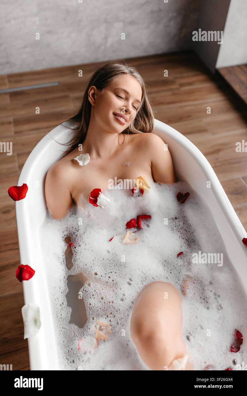 Giovane donna che si gode un bagno rilassante con schiuma e petali di fiori, concetto di spa Foto Stock
