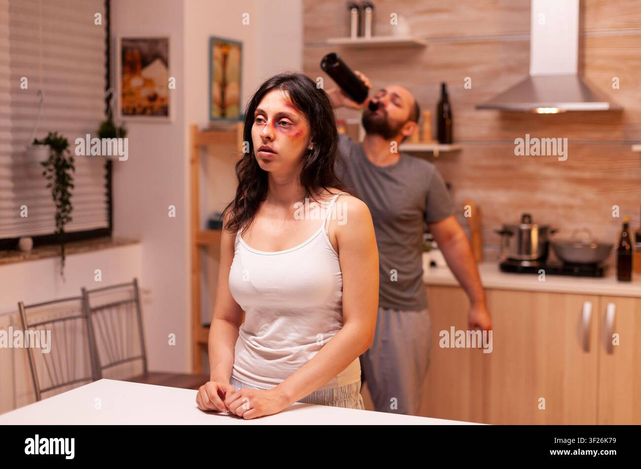 Marito violento beve alcol dopo aver picchiato la moglie ferita, mostrando abusi domestici, paura e l'impatto del comportamento aggressivo a casa. Foto Stock