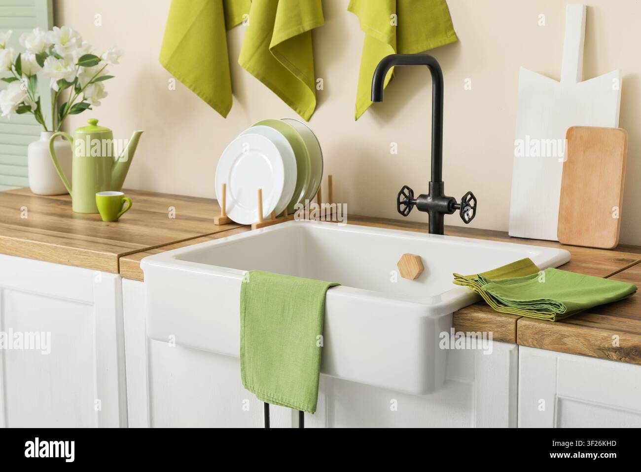Interno di cucina moderna con lavello stile fattoria bianco, rubinetto nero, piano di lavoro in legno e accenti verdi Foto Stock