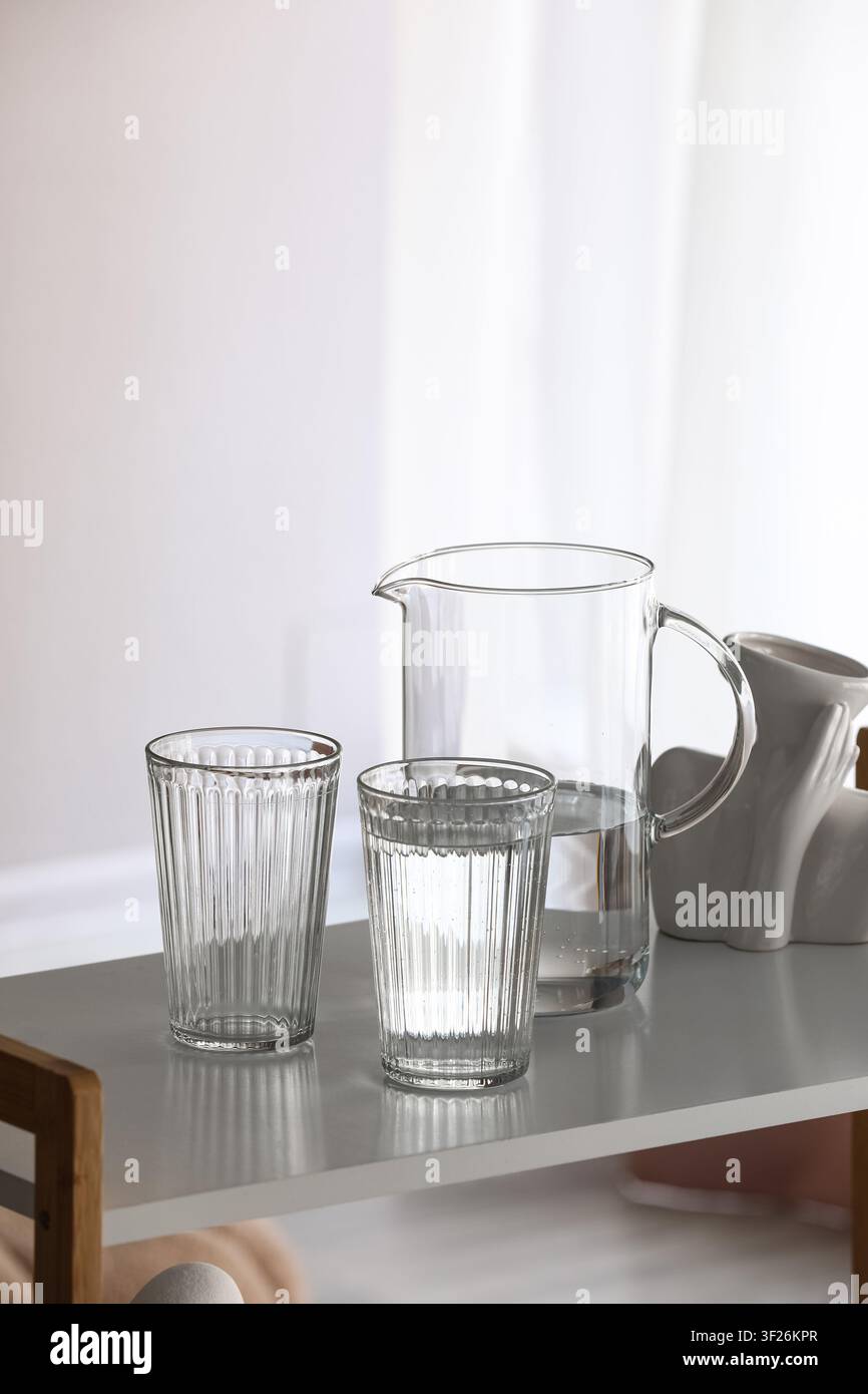 Due bicchieri di vetro rigati e una caraffa di vetro trasparente con acqua su uno scaffale bianco, luce naturale e ombre Foto Stock