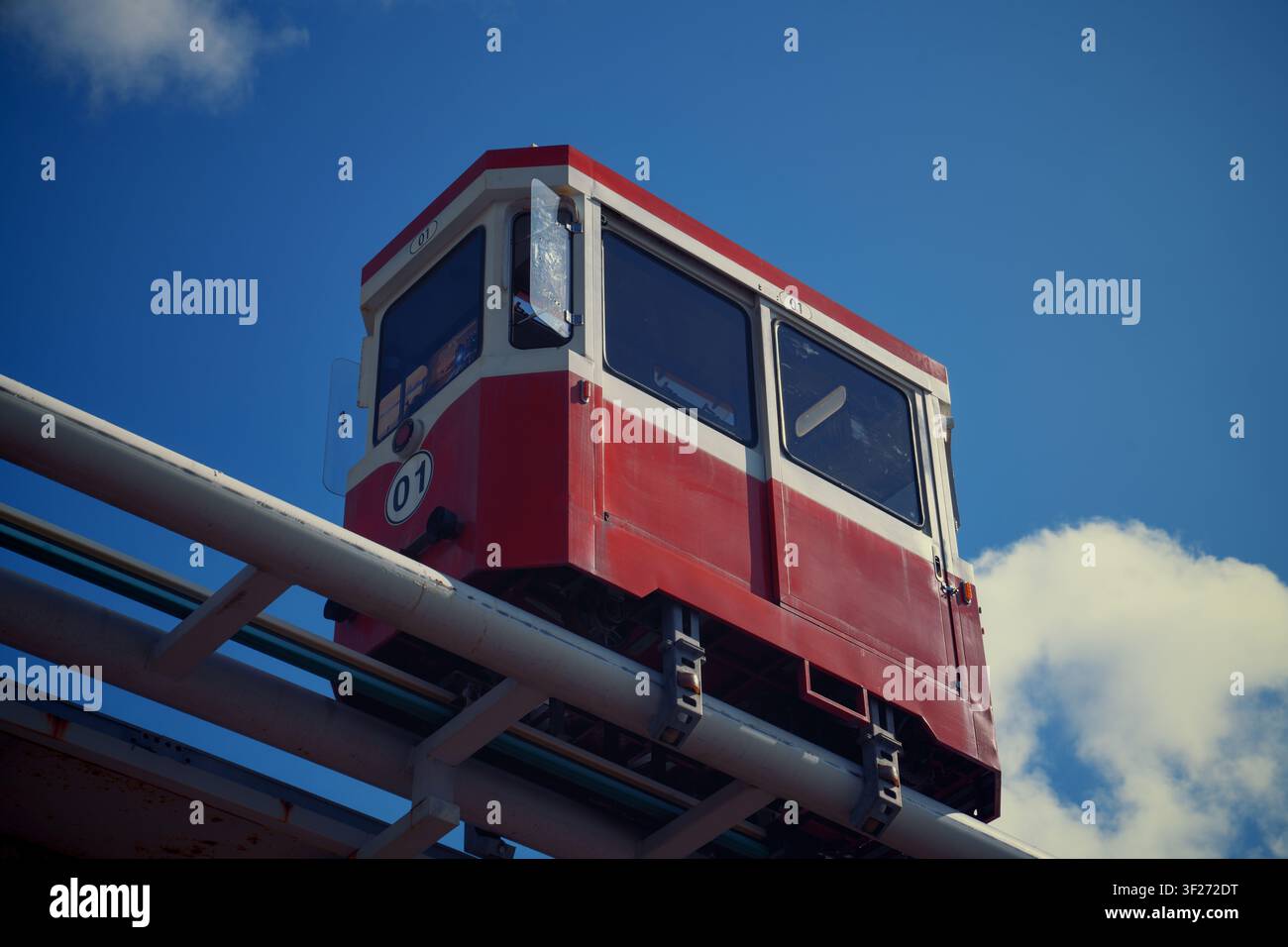 Una capsula rossa vibrante che si muove su una guida su elevata in monorail metallico contro un cielo blu profondo con nuvole soffici. Foto Stock