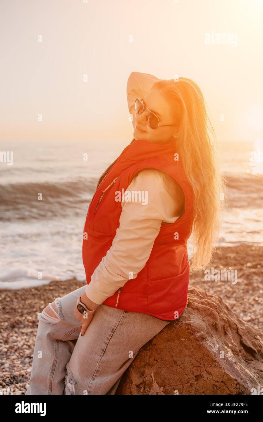 Donna sulla spiaggia al tramonto, seduta su una roccia vicino all'oceano durante l'ora dorata, godendosi la luce calda e la vista scenografica Foto Stock