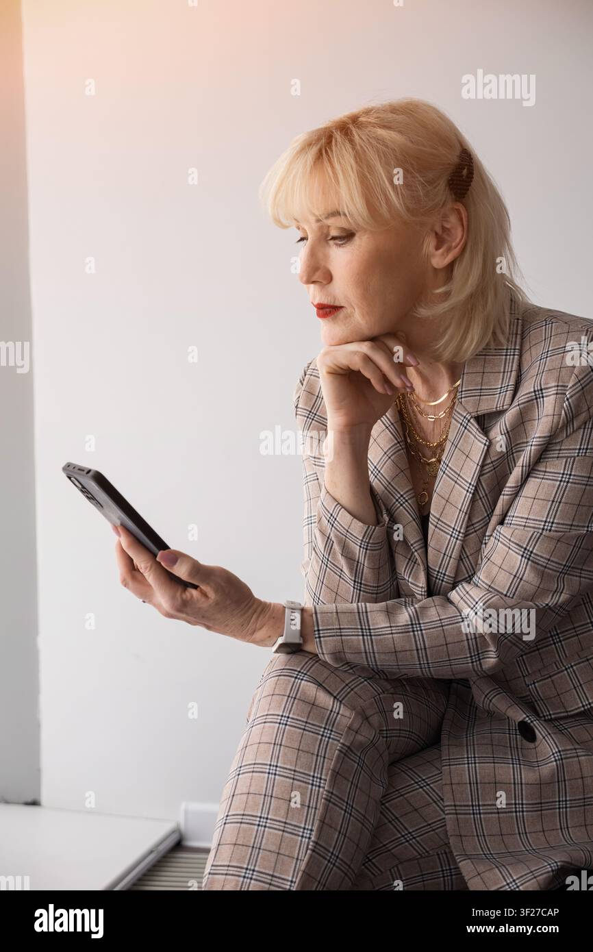 Donna d'affari bionda anziana in un completo a quadri è seduta al chiuso, concentrata sul suo smartphone, sfogliando pensierosa contenuti digitali ed email, trasmette Foto Stock