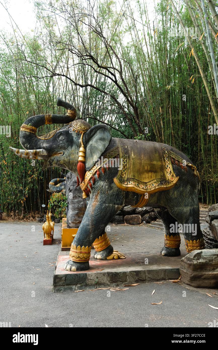 Statua decorativa di elefante con trunk sollevato e decorazioni dorate in un giardino di bambù, poggiata su una base di pietra in un tranquillo contesto di parco all'aperto Foto Stock