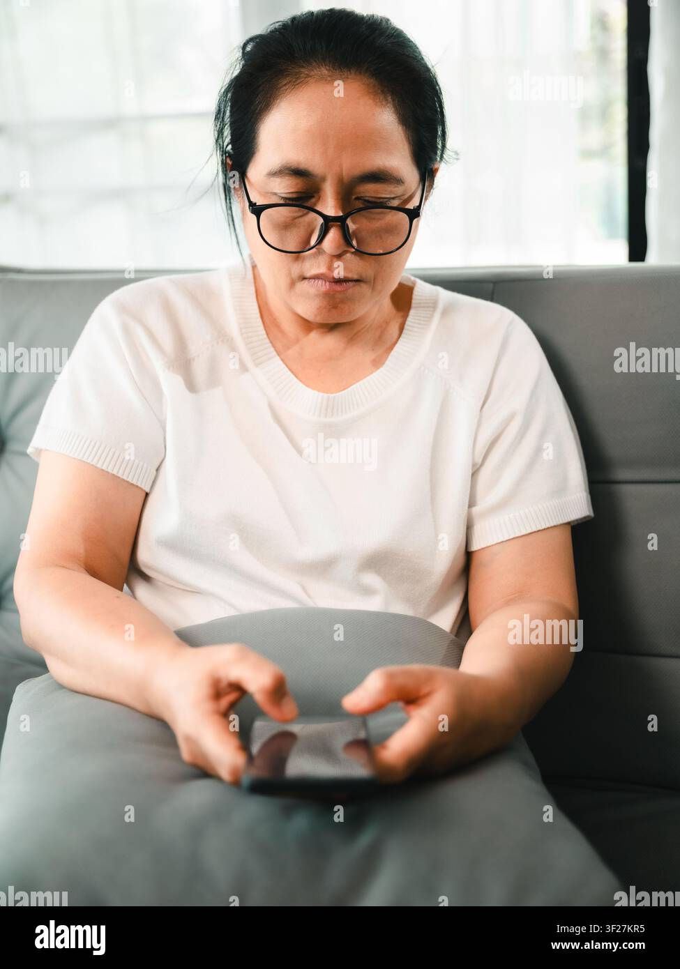 Donna asiatica di mezza età con occhiali che usa lo smartphone sul divano a casa, concetto di banking online, app mobile, comunicazione digitale, telemedicina, un Foto Stock