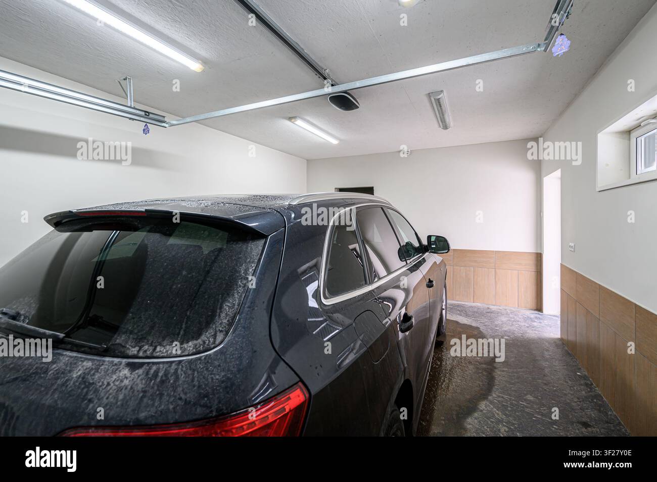 Un SUV scuro è parcheggiato in un garage con pareti e soffitto bianchi e dettagli in piastrelle. Il pavimento è bagnato e luci illuminano lo spazio Foto Stock