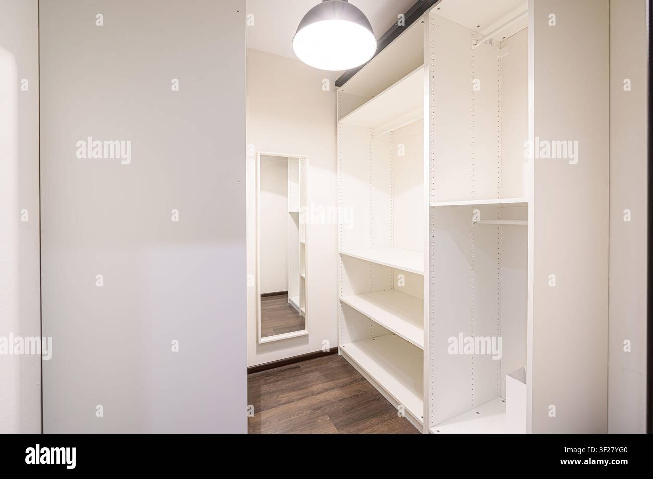 Viene mostrato un armadio a muro con scaffali bianchi, uno specchio a figura intera e una lampada a sospensione. Il pavimento è in legno scuro Foto Stock