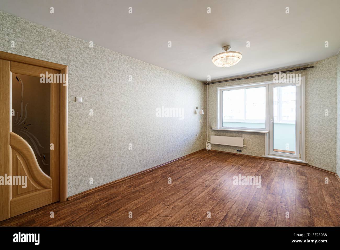 stanza luminosa e vuota con pavimenti in legno, carta da parati a motivi e una porta che conduce a un balcone. Illuminazione semplice e radiatore visibile Foto Stock